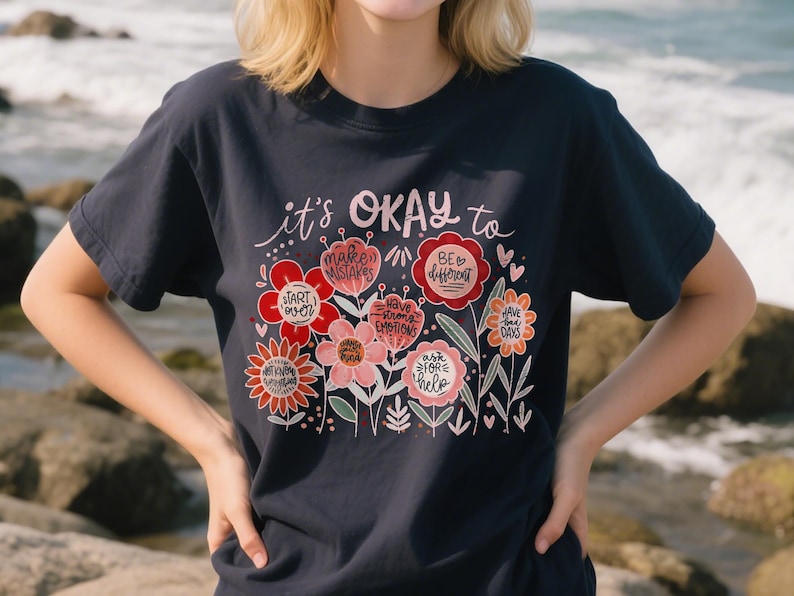 Op de afbeelding: Marineblauwe t-shirt met een bloemenontwerp en de tekst "it's OKAY to" boven de bloemen. De bloemen zijn in tinten rood, roze en oranje, met zinnen als "make mistakes" en "be different".