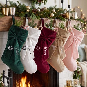 May include: Five velvet Christmas stockings in dark green, white, deep red, tan, and light pink. Each stocking features a large fabric bow on the front, a personalized name embroidered in white, and a small embroidered icon of an elf, tree, gingerbread man, holly, or snowflake.