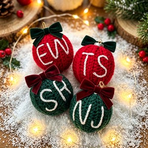 May include: Four round, crocheted Christmas ornaments in red and dark green. Each ornament features a dark velvet bow at the top and white embroidered letters on the front. The letters on the ornaments spell out EW, TS, SH, and NJ respectively.