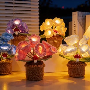 Puede incluir: Cinco lámparas de flores de ganchillo en azul, rosa, amarillo, blanco y morado. Cada lámpara presenta flores brillantes, hojas verdes y un acento de hongo, todo en una maceta tejida marrón. Las lámparas están iluminadas.