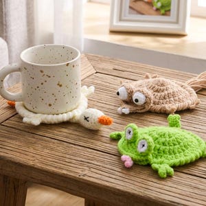 以下が含まれることがあります： 茶色の斑点があるクリーム色の陶器製マグカップが、白いかぎ針編みのカモのコースターの上に置かれています。その横には、大きな目をしたベージュの生き物の形をしたコースターと、鮮やかな緑色のカエルの形をしたコースターの2つのかぎ針編みのコースターがあります。