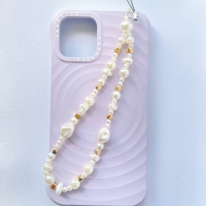 Op de afbeelding: Een lichtpaarse telefoonhoes met een telefoonriem van parels en gouden kralen. De riem bestaat uit witte parels en kleine gouden kralen. De telefoonhoes heeft een gladde, golfachtige textuur.