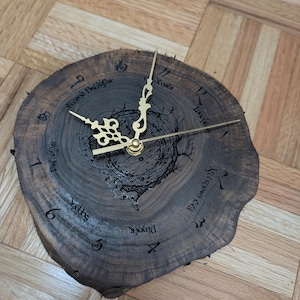 Op de afbeelding: Een ronde houten klok met een donkerbruine afwerking. De wijzerplaat is gemaakt van een dwarsdoorsnede van een boomstam, met goudkleurige wijzers en Romeinse cijfers. De klok heeft een uniek, rustiek ontwerp.