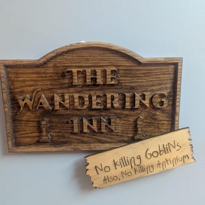 Puede incluir: Un letrero de madera con las palabras "THE WANDERING INN" talladas. Debajo del letrero hay una placa de madera más pequeña que dice "No killing Goblins. Also, No killing Antinium."