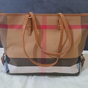 Könnte beinhalten: Eine beigefarbene Tragetasche mit einem klassischen Karomuster in Braun-, Rot- und Weißtönen. Die Tasche hat braune Ledergriffe und -verzierungen. Die Tasche hat eine rechteckige Form und einen geräumigen Innenraum.