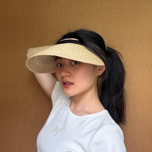 May include: A straw visor with a black band, worn by a person. The visor has a wide brim and is made of woven straw material. The person is wearing a white t-shirt.