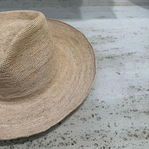 May include: A beige woven sun hat with a wide brim. The hat is made of a natural fiber material and has a rounded crown. The brim is approximately 5 inches wide. The hat is sitting on a light gray surface.