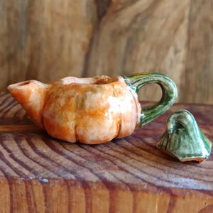 Può includere: Piccola teiera in ceramica a forma di zucca, corpo arancione e bianco, manico verde. Il coperchio verde è separato. La teiera è appoggiata su una superficie di legno.