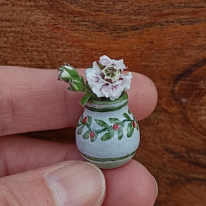 Könnte beinhalten: Eine kleine Keramikvase mit hellblauem Grund, verziert mit grünen Streifen und roten Beerenmotiven. Die Vase enthält eine zarte weiß-violette Blume mit grünen Blättern und einer Knospe. Die Vase wird in einer Hand gehalten.