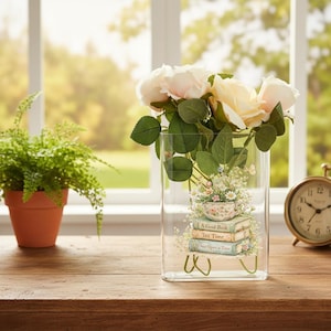 May include: A clear glass vase with a floral design and a bouquet of cream-colored roses. The vase has a decorative image of a teacup, books, and the words "A Good Book", "Tea Time", and "Once Upon a Time".