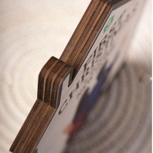 May include: Wooden sign with the text "FIRST CHRISTMAS" in a decorative font. The sign has a dark brown, layered wood edge and a white face with colorful accents. The sign is angled, showing the side and front.