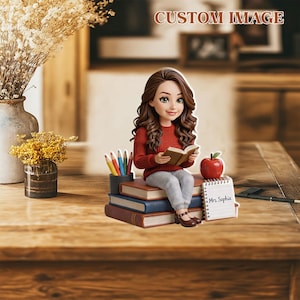 Puede incluir: Una figura de una mujer con cabello castaño largo, vestida con un suéter rojo y pantalones grises, leyendo un libro mientras está sentada en una pila de libros. También hay una manzana, lápices y un bloc de notas con el nombre "Mrs. Sophia". Las palabras "CUSTOM IMAGE" están en la parte superior.