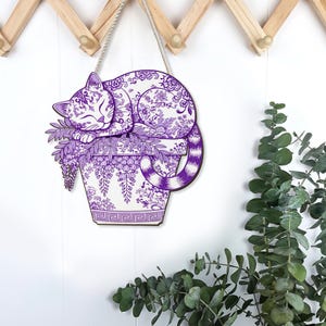 May include: A purple and white cat-shaped wall hanging. The cat is curled up on a flower pot with a floral design. The cat and pot have a detailed pattern. The wall hanging is suspended by a white rope.