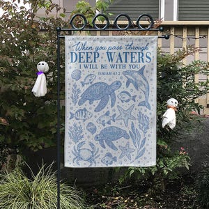 Op de afbeelding: Een blauw-witte tuinvaandel met illustraties van zeedieren, waaronder schildpadden, vissen en een dolfijn. De vlag toont de tekst "When you pass through deep waters, I will be with you" en "Jesaja 43:2".
