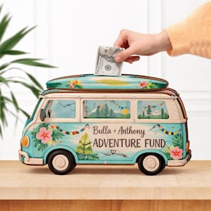 May include: A teal and cream-colored camper van-shaped bank with a surfboard on top. The bank has floral and landscape designs, with the text "Bulla + Anthony ADVENTURE FUND". A hand is inserting a $100 bill into the slot.