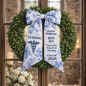 May include: A green wreath with a large white and blue floral bow. The bow has the text "NURSING" and "Jessica Williams BSN RN University School of Nursing Class of 2026". The wreath is hanging on a wooden window.