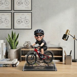 Peut inclure: Figurine personnalisée d'une personne sur un VTT rouge. La figurine est posée sur un socle avec le nom "ANTHONY". Sur un bureau avec une lampe, des livres et des impressions encadrées de brevets de vélos.