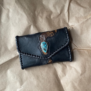 Puede incluir: Una cartera de cuero negro con una incrustación de piel de serpiente marrón texturizada en la solapa. La cartera está decorada con una piedra turquesa en forma de lágrima engastada en plata en el centro y rematada con costuras negras.