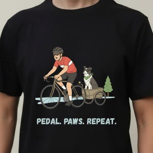 Peut inclure: T-shirt noir avec un cycliste et un chien dans une remorque. Le cycliste porte un maillot rouge et blanc et un casque. Le chien porte un bandana vert. Le texte "PEDAL. PAWS. REPEAT." est imprimé en dessous.