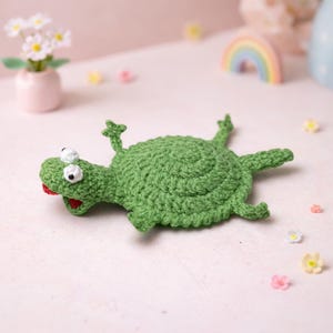 May include: A handmade green crochet turtle with white eyes and a red tongue. The turtle is lying on its back with its legs and head extended. The crochet work has a textured appearance.