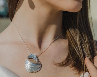 Puede incluir: Un collar de plata con un colgante grande y texturizado en forma de concha. También se ven pendientes a juego con una pequeña piedra verde rectangular. La joya se lleva sobre una parte superior blanca.