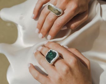 May include: Two silver rings with large, irregularly shaped gemstones. One ring features a light green stone, the other a dark green stone. The rings are set against a backdrop of white fabric and a blurred background.
