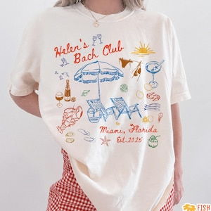 May include: Cream-colored t-shirt with a beach-themed graphic. The design includes an umbrella, beach chairs, and the text "Helen's Bach Club Miami, Florida Est. 2025". The shirt is paired with red and white checkered shorts.