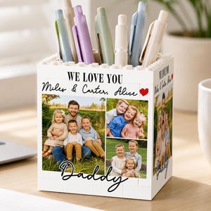 May include: White block-style pen holder with family photos and the text "WE LOVE YOU Miles & Carter, Alice Daddy". The holder contains several pens in various colors, including green, purple, and blue.