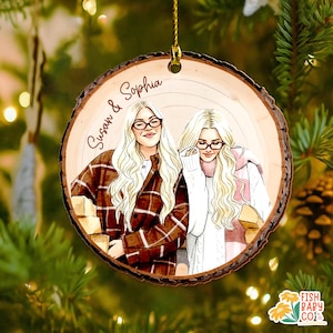 May include: A round wooden Christmas ornament with a hand-drawn illustration of two women with blonde hair and glasses. The ornament has the names "Susan & Sophia" written on it. The women are wearing sweaters and jackets. The ornament is hanging from a gold string.