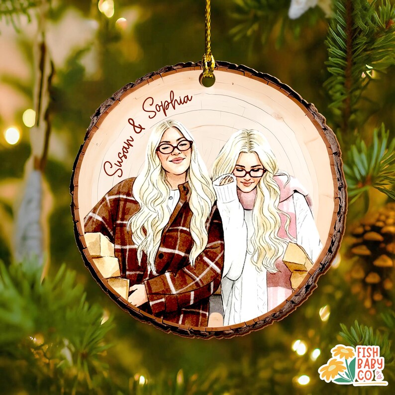 May include: A round wooden Christmas ornament with a hand-drawn illustration of two women with blonde hair and glasses. The ornament has the names "Susan & Sophia" written on it. The women are wearing sweaters and jackets. The ornament is hanging from a gold string.