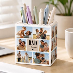 May include: A white pen holder with a photo collage of a father and children. The pen holder has a block design and the words "DAD we love you" are printed on the front. Various pens in pastel colors are in the holder.