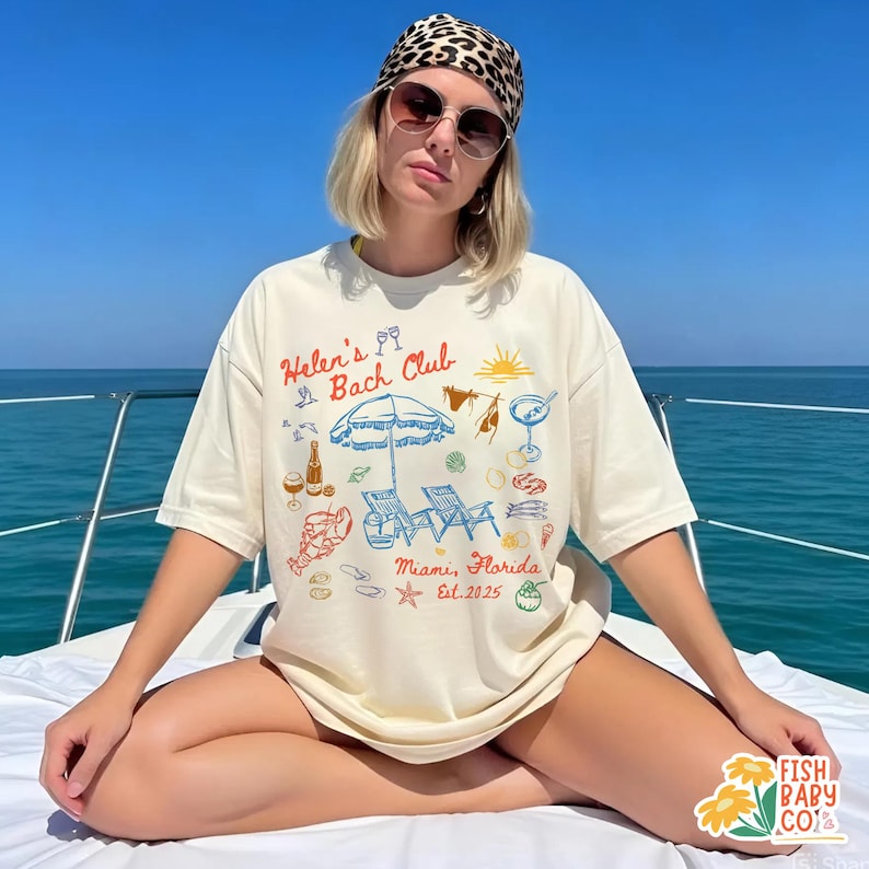 May include: A cream-colored short-sleeve t-shirt featuring a colorful graphic print with the text "Helen's Bach Club" and "Miami, Florida Est. 2025". The design includes illustrations of a beach umbrella, lounge chairs, a lobster, and drinks.