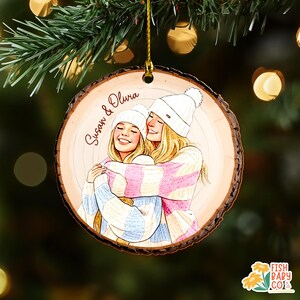 May include: A wooden Christmas ornament with a drawing of two people hugging, wearing white beanies and sweaters. The ornament has the names "Susan & Olivia" written on it. The ornament is hanging from a gold string.