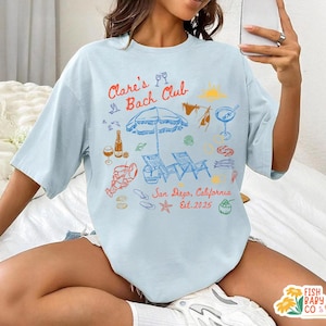 May include: A light blue t-shirt featuring a colorful graphic print with the text "Clare's Bach Club" and "San Diego, California Est. 2025". The design includes illustrations of a beach umbrella, lounge chairs, a lobster, drinks, and seashells.