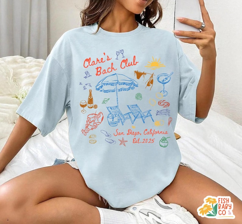 May include: A light blue t-shirt featuring a colorful graphic print with the text "Clare's Bach Club" and "San Diego, California Est. 2025". The design includes illustrations of a beach umbrella, lounge chairs, a lobster, drinks, and seashells.