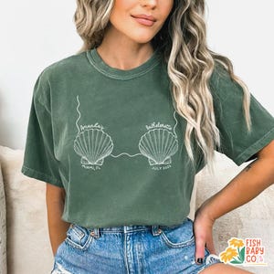 May include: Green t-shirt with a white seashell design. The text on the shirt reads "Amanda's Bachelorette" with "Miami, FL" and "July 2025" below the seashells. The shirt is a relaxed fit.