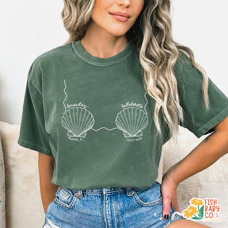 May include: Green t-shirt with a white seashell design. The text on the shirt reads "Amanda's Bachelorette" with "Miami, FL" and "July 2025" below the seashells. The shirt is a relaxed fit.