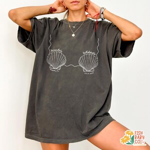 May include: Dark gray t-shirt with a white graphic of two seashells connected by a line, with the text "Amanda's Bachelorette" above and "Miami, FL July 2023" below. The shirt is a relaxed fit.