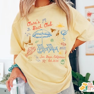 May include: A pale yellow t-shirt featuring a colorful graphic print with the text Clare's Bach Club, San Diego, California, Est. 2025. The design includes illustrations of a beach umbrella, lounge chairs, a lobster, drinks, and seashells.