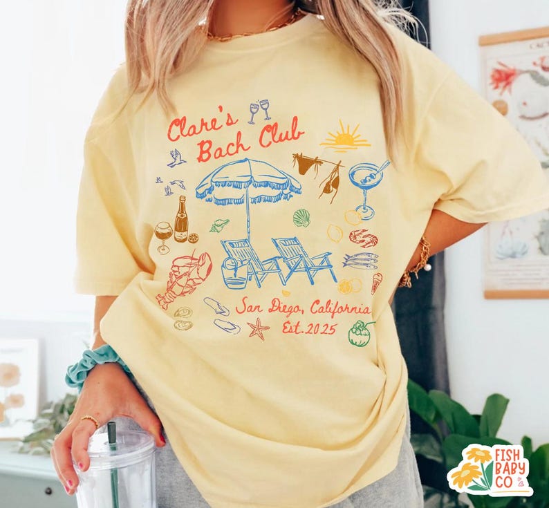 May include: A pale yellow t-shirt featuring a colorful graphic print with the text Clare's Bach Club, San Diego, California, Est. 2025. The design includes illustrations of a beach umbrella, lounge chairs, a lobster, drinks, and seashells.