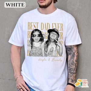 Puede incluir: Camiseta blanca con el texto dorado "BEST DAD EVER". La camiseta presenta una foto en blanco y negro de dos ni&ntilde;as, con los nombres "Kayla & Beverly" debajo. La camiseta tiene cuello redondo.