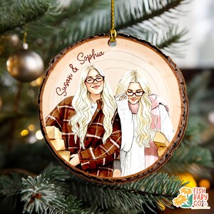 May include: A wooden Christmas ornament featuring a hand-drawn illustration of two women with blonde hair and glasses. The ornament has the names "Susan & Sophia" written on it. The women are wearing sweaters and jackets. The ornament is hanging from a gold string.