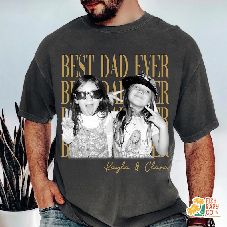 Puede incluir: Camiseta gris oscuro con una foto en blanco y negro de dos ni&ntilde;as, con el texto dorado "BEST DAD EVER". Los nombres "Kayla & Clara" est&aacute;n impresos debajo. La camiseta tiene un estilo vintage.