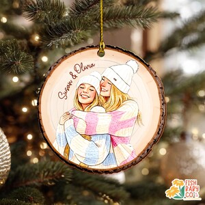 May include: A wooden Christmas ornament with a drawing of two people hugging, wearing white beanies and striped sweaters in pink and blue. The ornament has the names "Susan & Olivia" written in cursive. The ornament is hanging from a gold string.