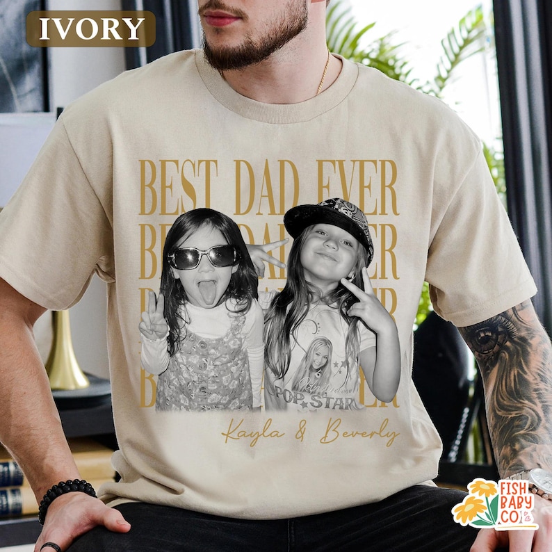 Puede incluir: Camiseta de color marfil con el texto dorado "BEST DAD EVER". Una foto en blanco y negro de dos ni&ntilde;as est&aacute; impresa en la camiseta, con los nombres "Kayla & Beverly" debajo. La camiseta es de estilo casual.