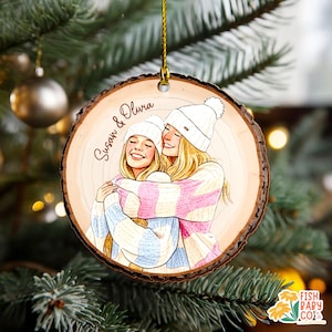 May include: A wooden Christmas ornament featuring a drawing of two people hugging, wearing white beanies and sweaters. The ornament has the names "Susan & Olivia" written on it. The ornament is hanging from a gold string.