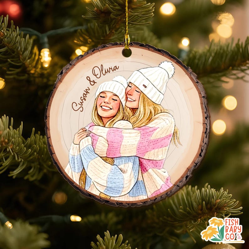 May include: A round wooden Christmas ornament with a drawing of two people hugging, wearing white beanies and striped sweaters. The ornament has the names "Susan & Olivia" written on it. The ornament is hanging on a Christmas tree.