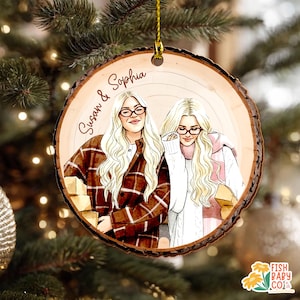 May include: A round wooden Christmas ornament with a drawing of two women with blonde hair and glasses. The ornament has the names "Susan & Sophia" written on it. The women are wearing sweaters and jackets. The ornament is hanging from a gold string.