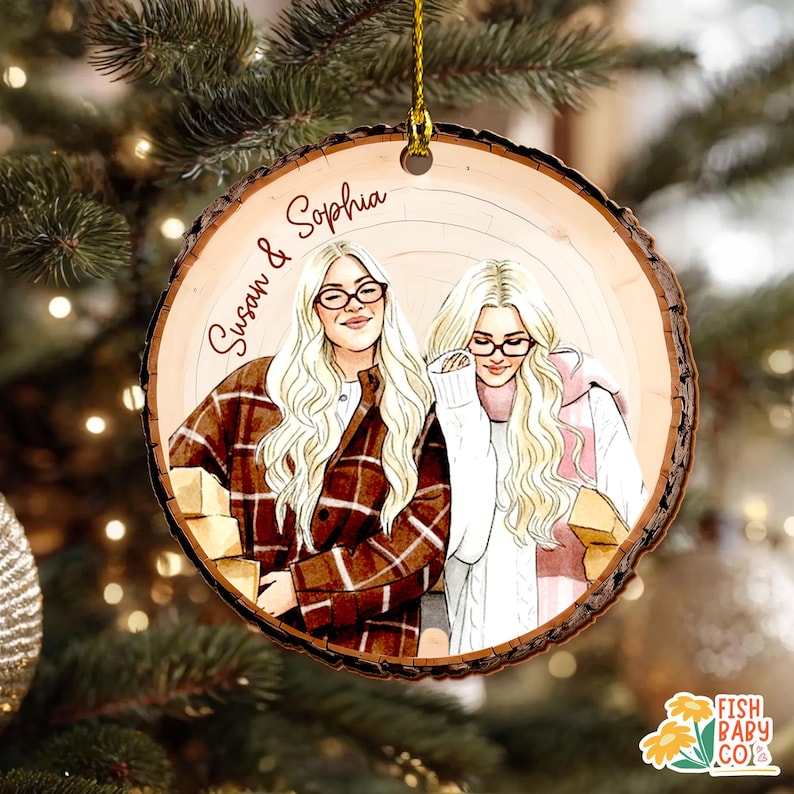 May include: A round wooden Christmas ornament with a drawing of two women with blonde hair and glasses. The ornament has the names "Susan & Sophia" written on it. The women are wearing sweaters and jackets. The ornament is hanging from a gold string.
