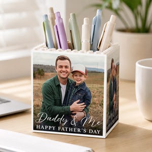 May include: A white pen holder with a photo of a man and child, with the text "Daddy & Me HAPPY FATHER'S DAY." The holder is filled with various colored pens. A laptop and a white mug are in the background.